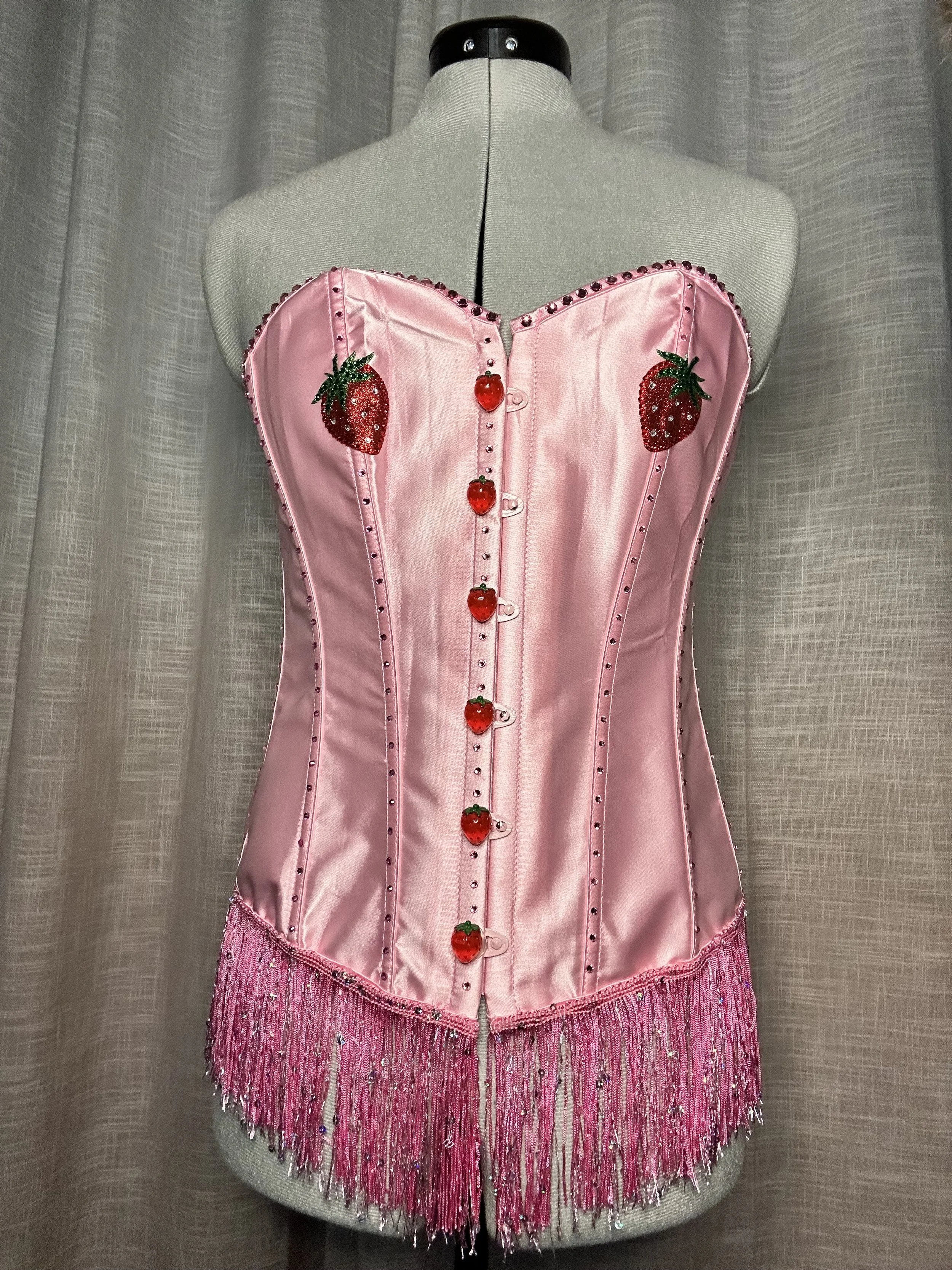 Pink satin bustier with strawberry and strawberry seed embellishments, buttons resembling strawberries, adorned with pink fringe and sparkly beads, displayed on a mannequin.