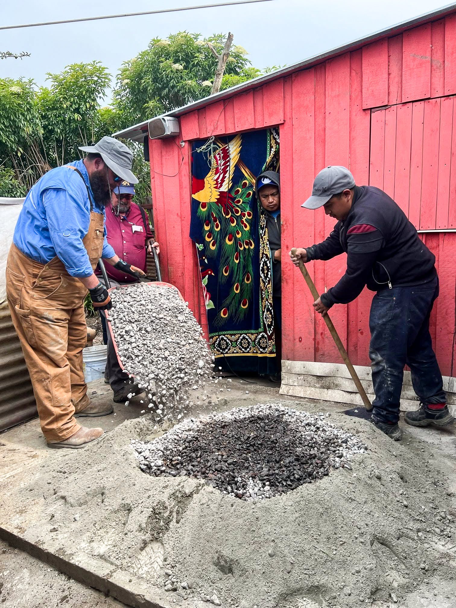 People working on a construction site, pouring and leveling concrete for a foundation or sidewalk outside a red building with a colorful patterned curtain.