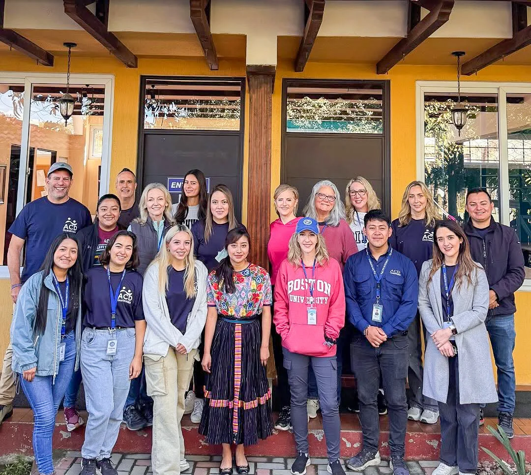 Group of diverse young adults and older women standing outside a yellow building with large windows, some with conference badges, smiling for a group photo