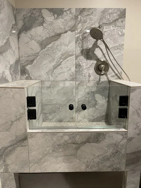 A shower area enclosed with a glass partition, featuring marble tiles on the wall and a handheld showerhead. The shower has black control knobs and two built-in shelves.