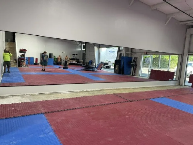 View of a martial arts gym with red and blue mats on the floor, a large mirror on the wall, and several people practicing or training.