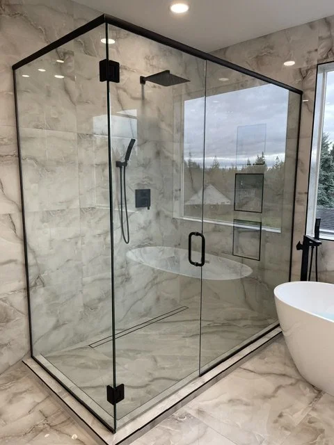 Modern glass shower enclosure in a bathroom with marble walls and floor, featuring a rainfall showerhead, hand-held shower, and built-in wall niches, with a freestanding bathtub visible nearby.