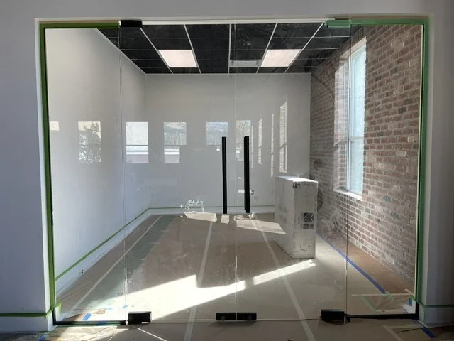 Empty office room with glass walls and door, brick wall on right, three windows, and construction materials on floor.