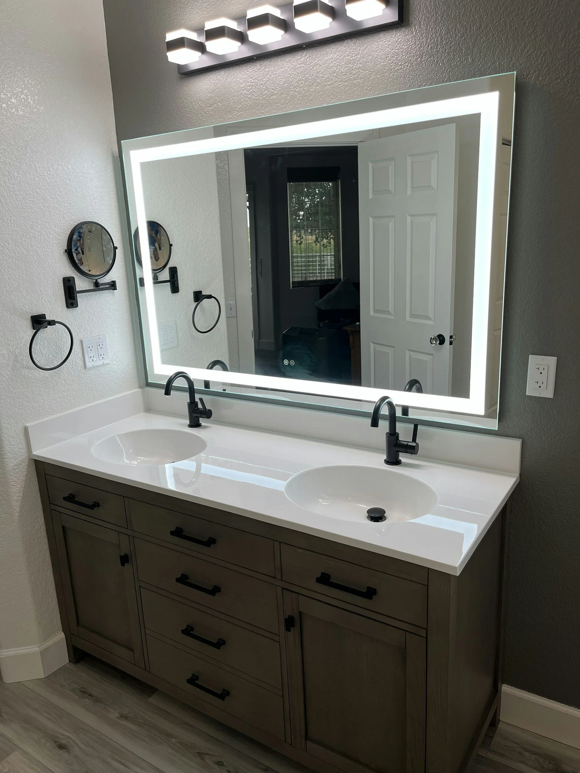 Modern bathroom vanity with dual sinks, a large backlit mirror, black fixtures, and a wooden cabinet with multiple drawers and handles, located in a room with textured gray walls and light-colored wood flooring.