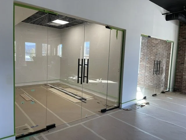 Office space under construction with glass walls and doors, exposed brick wall, and construction materials on the floor.