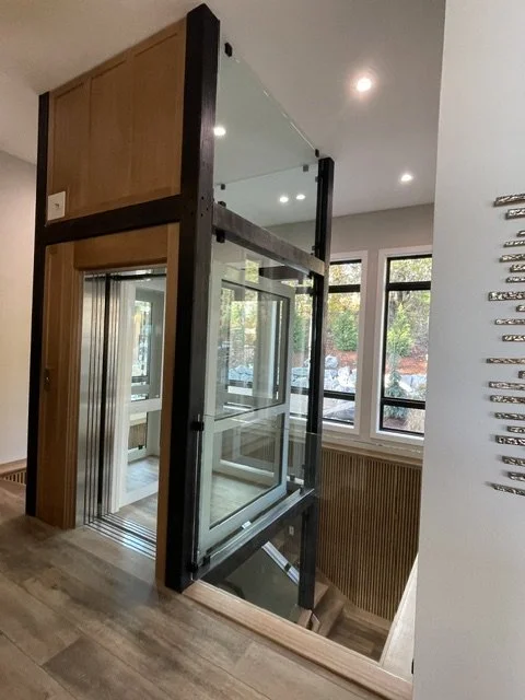 Modern interior with a glass elevator enclosed in black metal framework near windows.