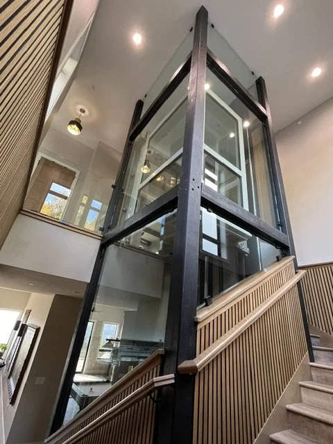 Interior view of a modern building with a glass elevator enclosed in a black metal frame, positioned next to a staircase with wooden handrails.