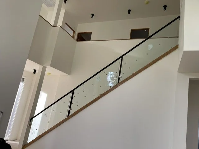 Interior view of a modern house staircase with glass railing and wooden handrail, leading to the upper floor with three doorways visible.