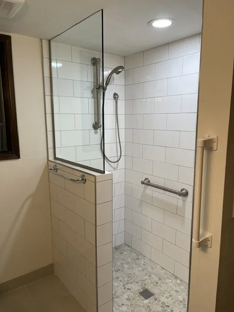 Accessible shower area with beige tiled walls, a glass partition, a handheld showerhead, and a grab bar.