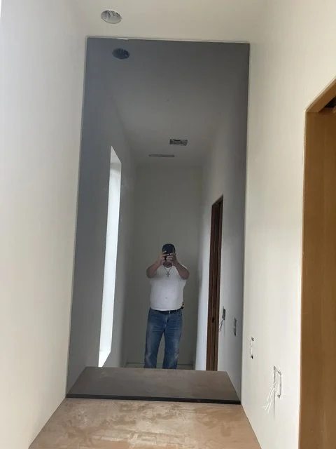 A person taking a photo of a tall mirror in a hallway, wearing a white shirt and blue jeans, with a doorway visible on the right side.