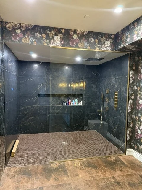 Empty walk-in shower with black marble walls, a built-in niche with toiletries, and floral wallpapered ceiling and surrounding walls.