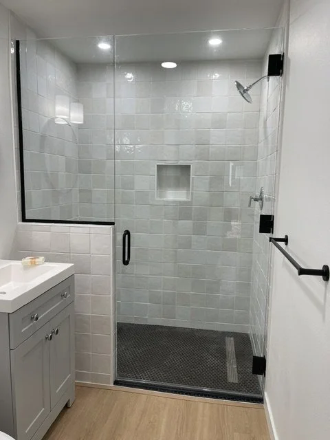 Glass shower enclosure with white tiled walls, a built-in niche, showerhead, and a black grab bar in a bathroom with a wooden floor and white vanity with a towel bar.