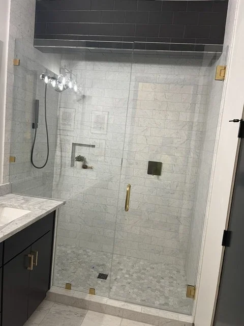 A modern bathroom shower with glass door, white tiled walls, and a built-in shelf with decorative items. There is a black cabinetry and marble countertop on the left side.