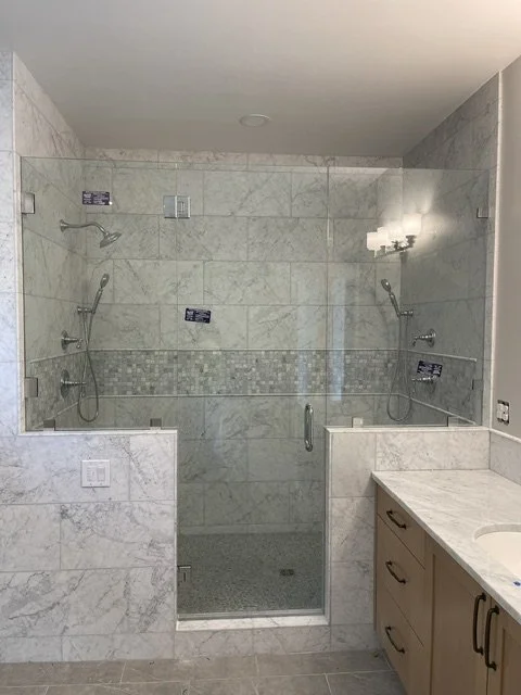 A bathroom shower area with two showerheads, glass enclosure, marble tiles, and a bathroom vanity with sink and cabinet.