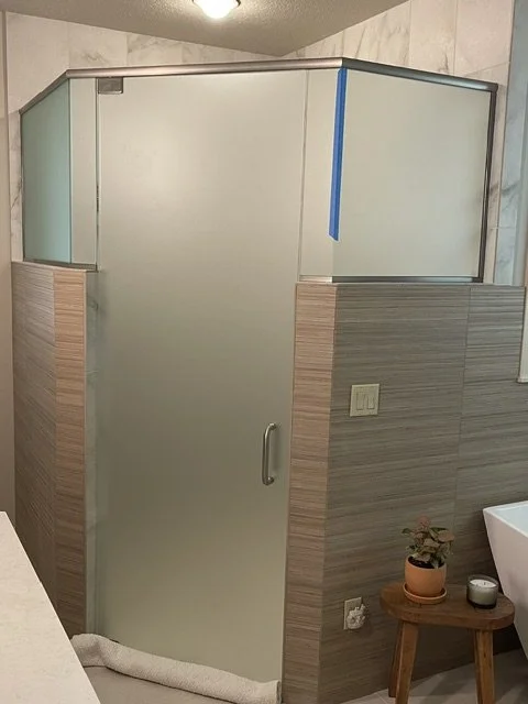 A bathroom shower with frosted glass door and walls, part of a tiled bathroom wall, a small wooden table with a potted plant and a candle, and an electrical outlet.