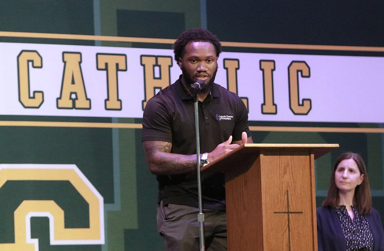 A man with dark curly hair and a beard speaking at a podium with a cross on it, wearing a black polo shirt, in front of a large banner with the word 'CATHOLIC' written on it; a woman with brown hair in a blue jacket is sitting to his left.