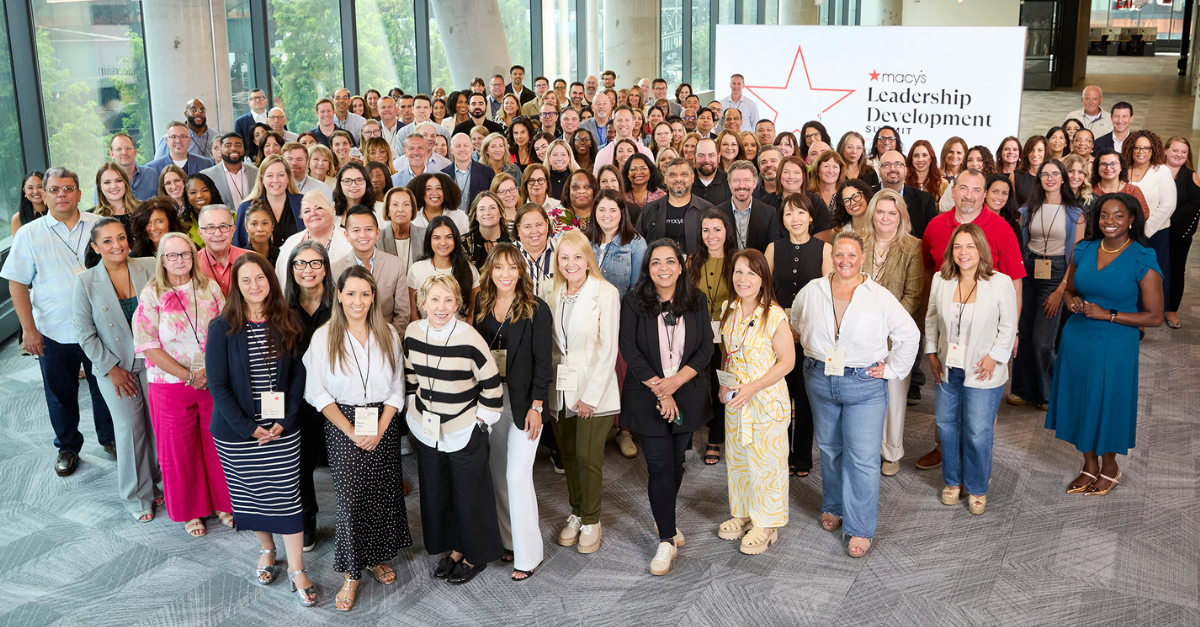 A large group of Macy's leaders posed together at the Leadership Development Summit, standing in a bright open room with a Macy's Leadership Development Summit banner behind them.