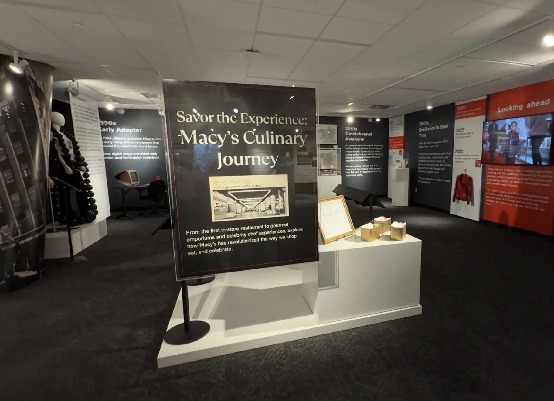 Interior of the Macy's heritage museum, a long gallery with timeline panels along the back wall marking the 1990s, 2010s, and 2020s into a "Looking ahead" section, display cases and a "Macy's Culinary Journey" banner in the foreground.