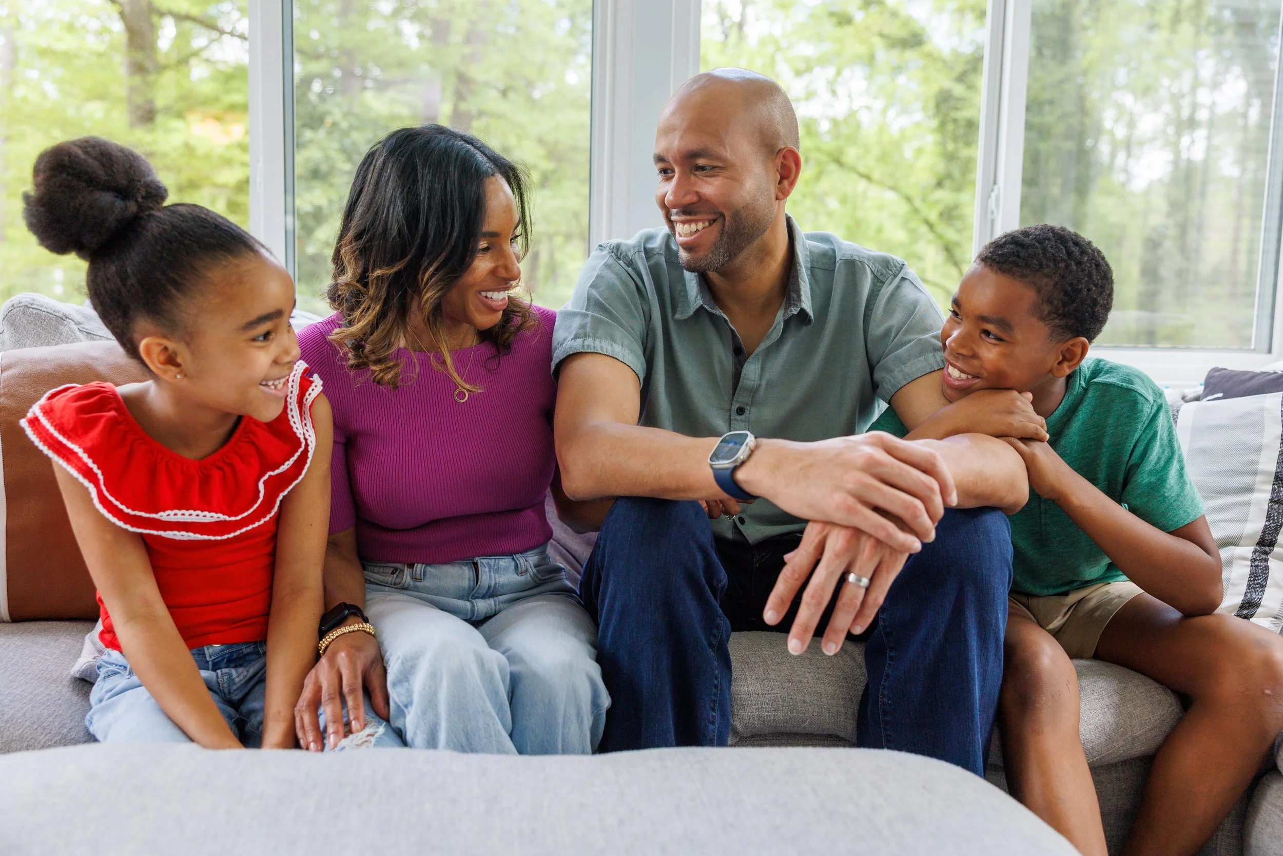 Jason Esteves, his wife Ariel, and their two children sitting on a couch in a bright living room with large windows, smiling and interacting with each other.