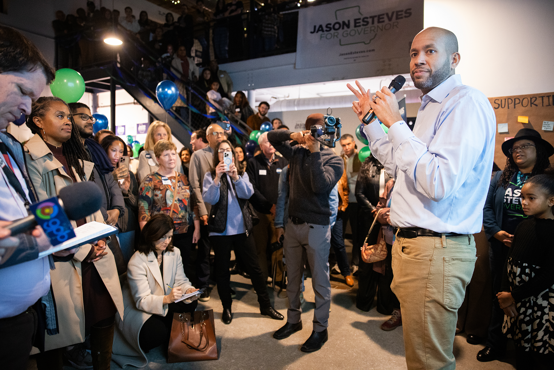 Jason Esteves addresses a large crowd at a political event with a Jason Esteves campaign banner in the background. The audience is listening attentively, some taking notes or photos, and there are balloons and a support sign on the wall.