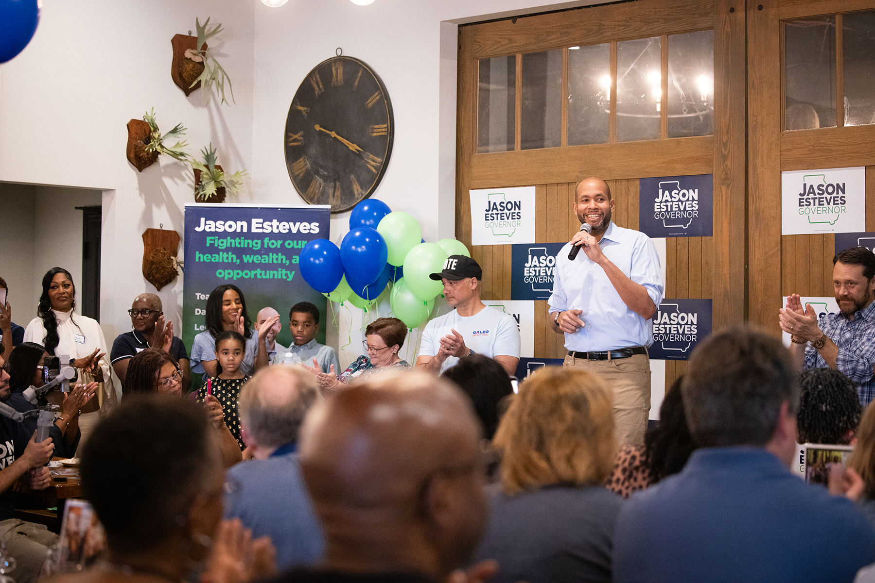 Georgia's Jason Esteves speaking into a microphone at a political rally surrounded by supporters, campaign signs for Jason Esteves for governor, and a backdrop with blue and green balloons.