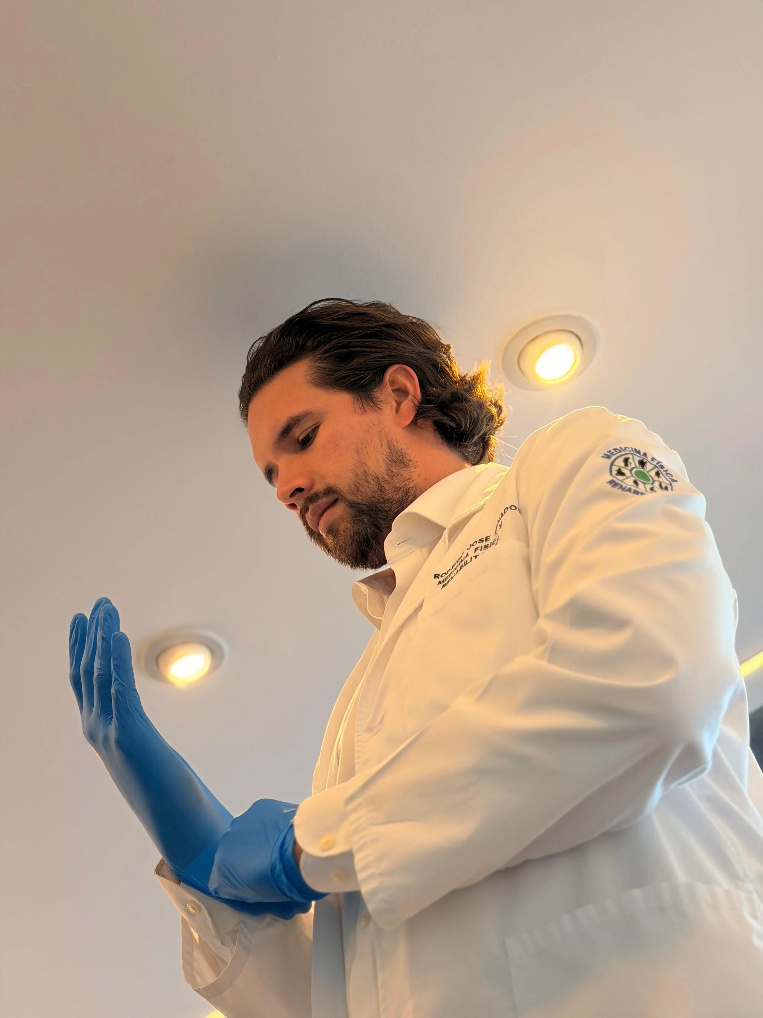 Un hombre con barba, traje de médico y guantes azules, con las manos levantadas en una actitud de saludo o oración, en un lugar con techo con luces empotradas.