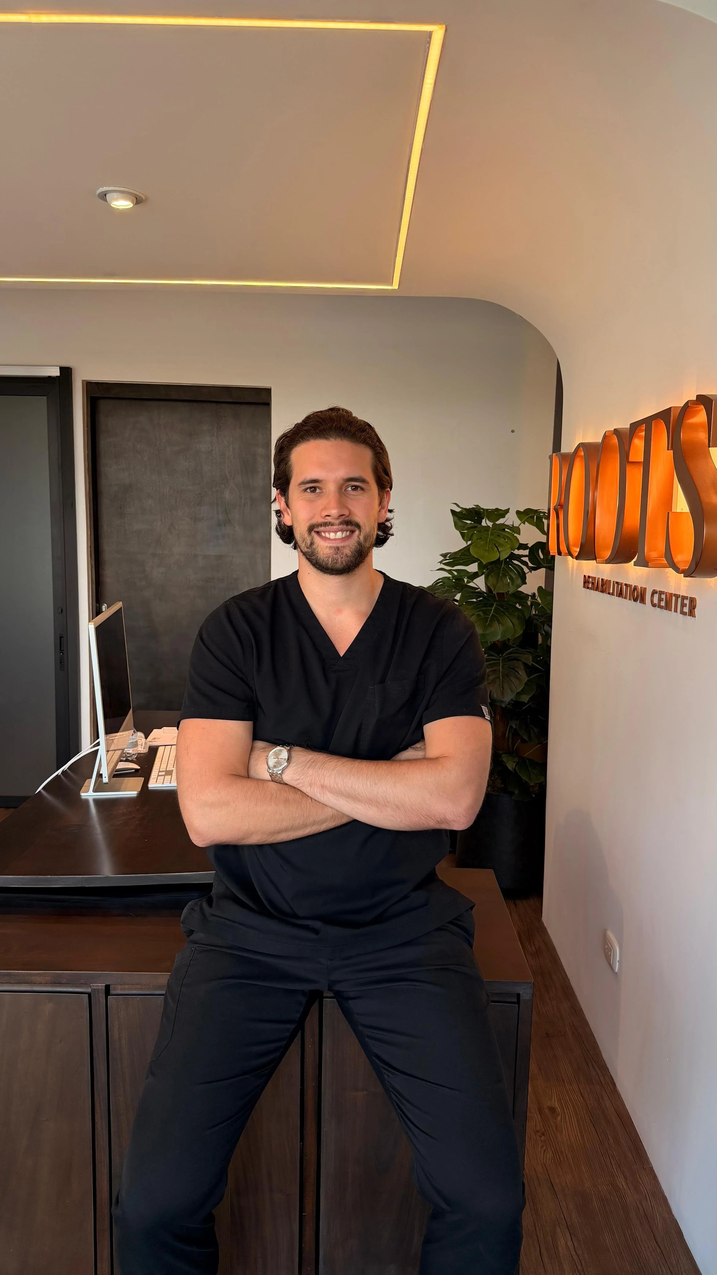 Hombre con barba y cabello oscuro, vestimenta médica negra, con brazos cruzados, posando en una clínica de rehabilitación, en un ambiente moderno con plantas y señal de 'Rehabilitation Center'