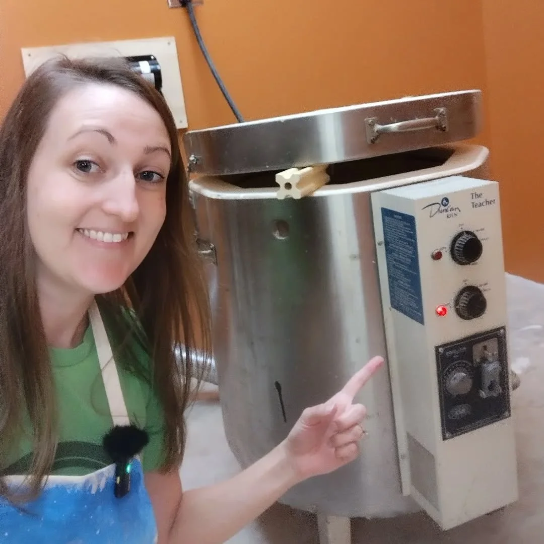 Woman smiling, pointing at a large stainless steel food steamer in a kitchen or food prep area.