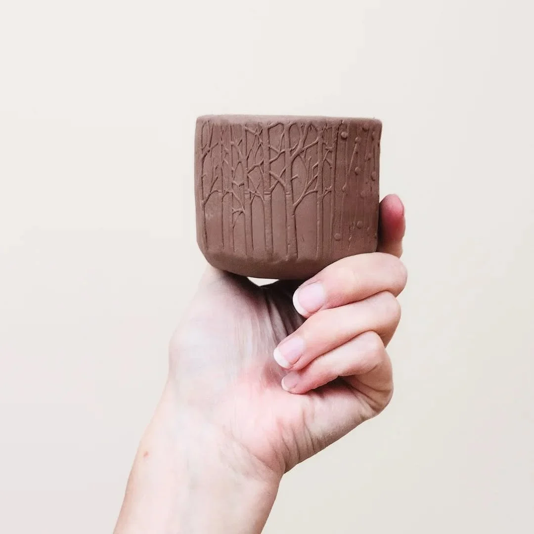 A hand holding a small clay cup with carved tree and branch designs on it.