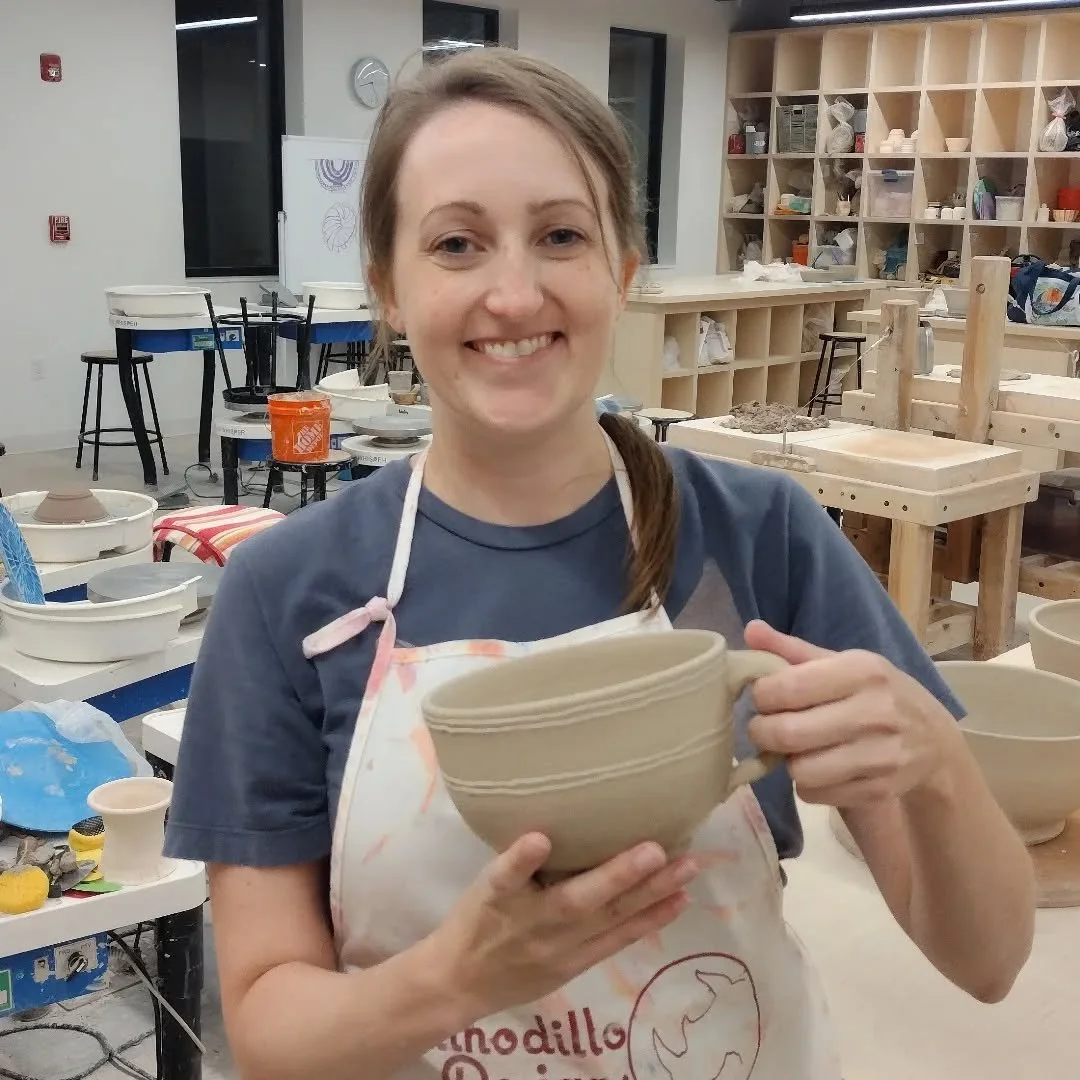 A woman smiling and holding a ceramic bowl in a pottery studio, surrounded by pottery tools and shelves with pottery supplies.