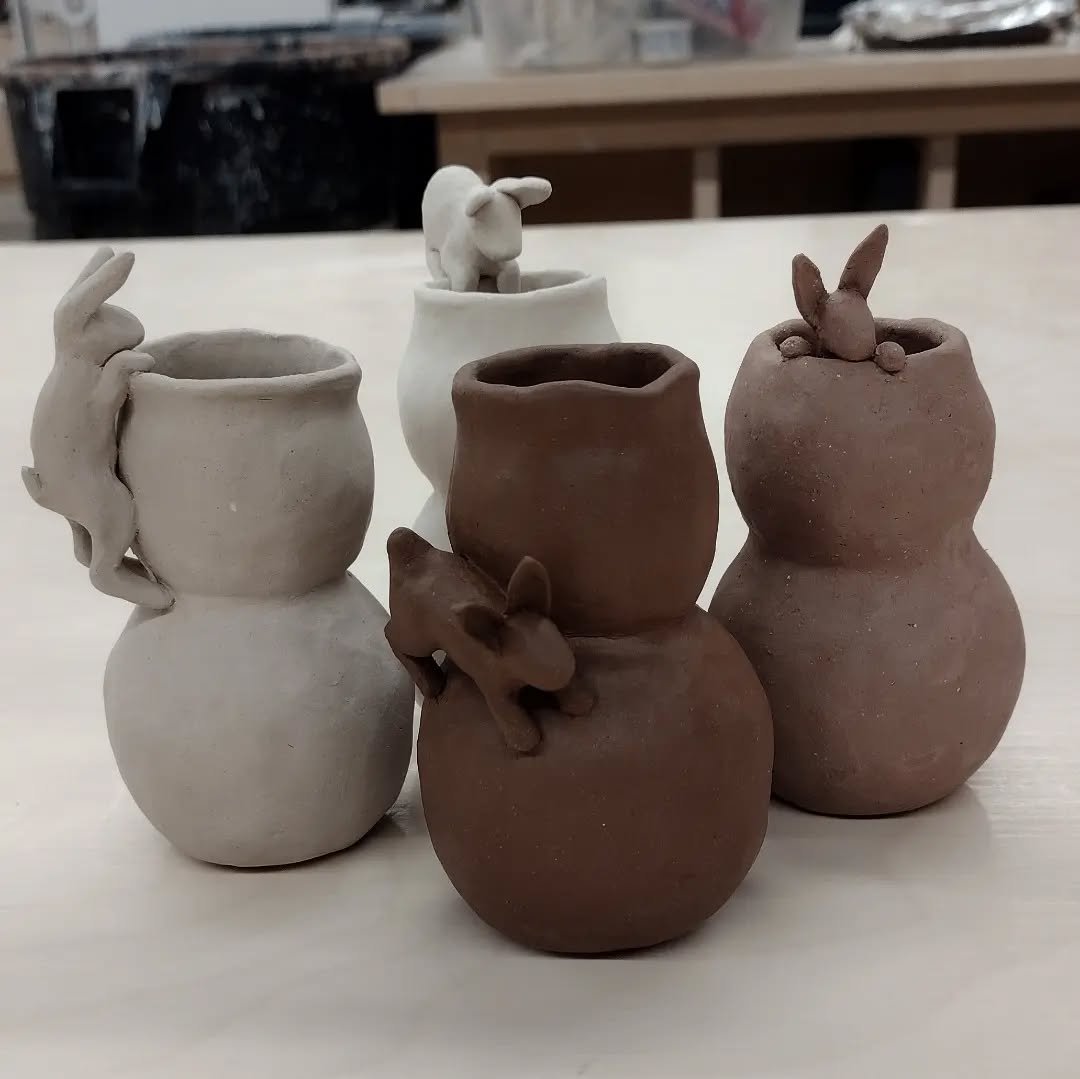 Six small clay vases with animal-shaped handles and decorations, including rabbits and a bear, displayed on a wooden surface.
