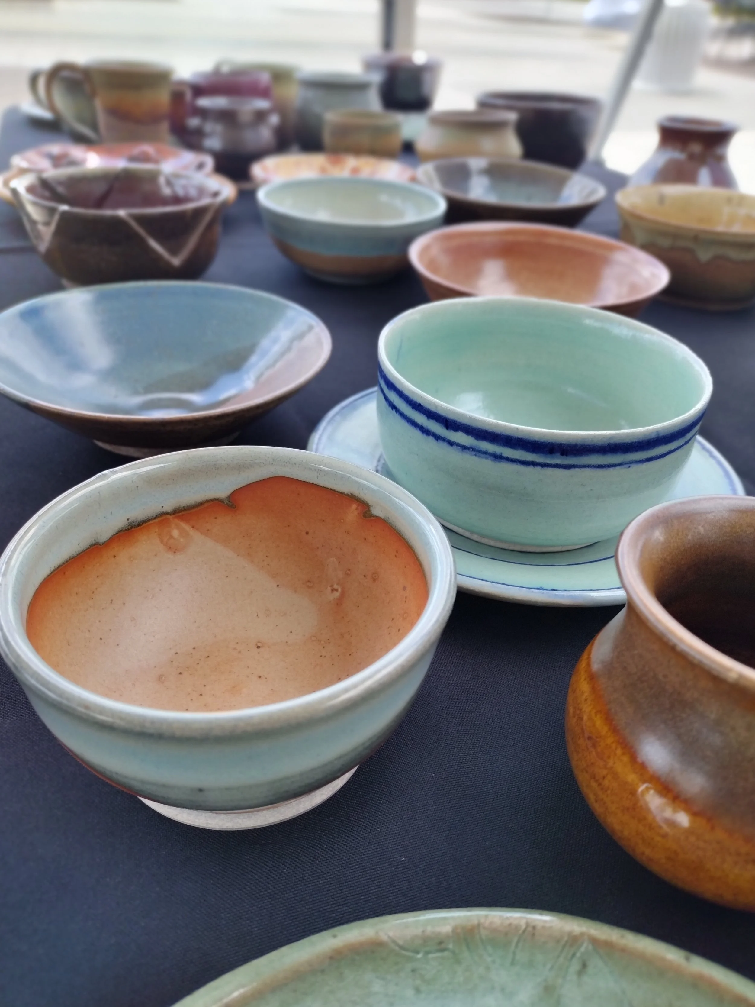 A display of colorful pottery bowls arranged on a table. The bowls are in various colors including green, brown, blue, and tan, and have different shapes and sizes.