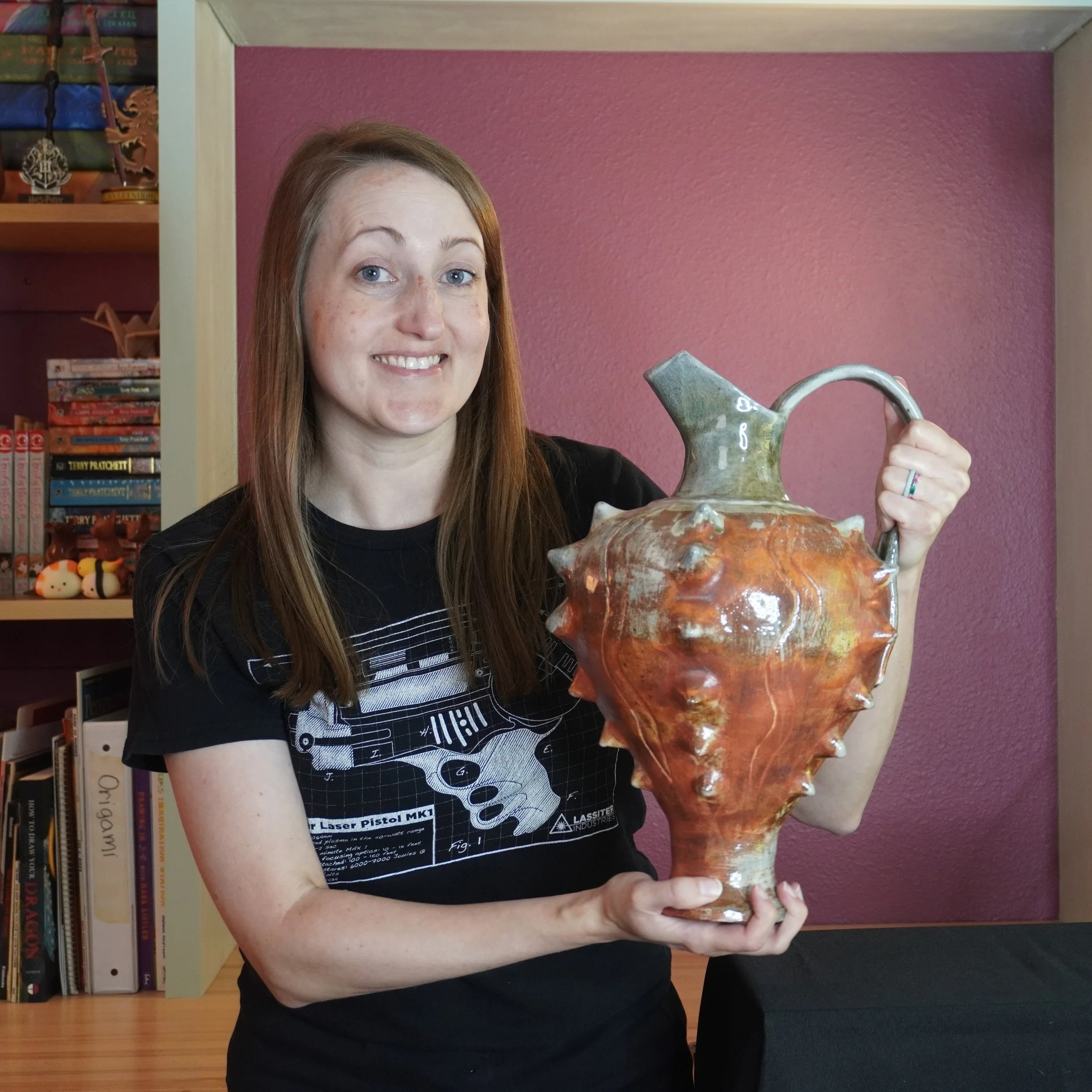 A woman with red hair and light skin, smiling, holding a large ceramic decorative vessel with a pointed top and rounded body, in front of a pink wall and bookshelf.