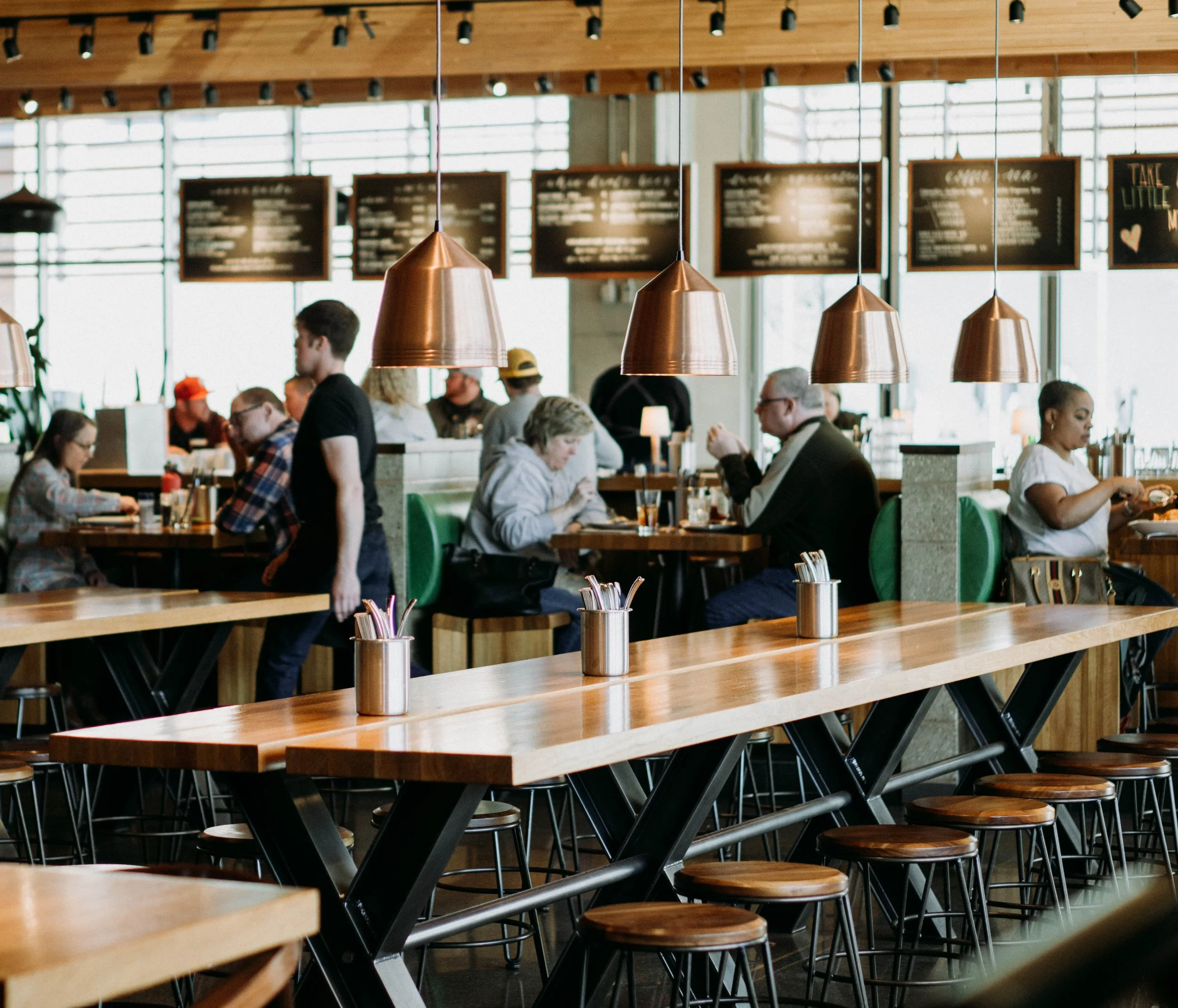 Interior of a modern restaurant or cafe with wooden tables, copper pendant lights, and large windows in the background. Customers are seated and interacting or using their devices.