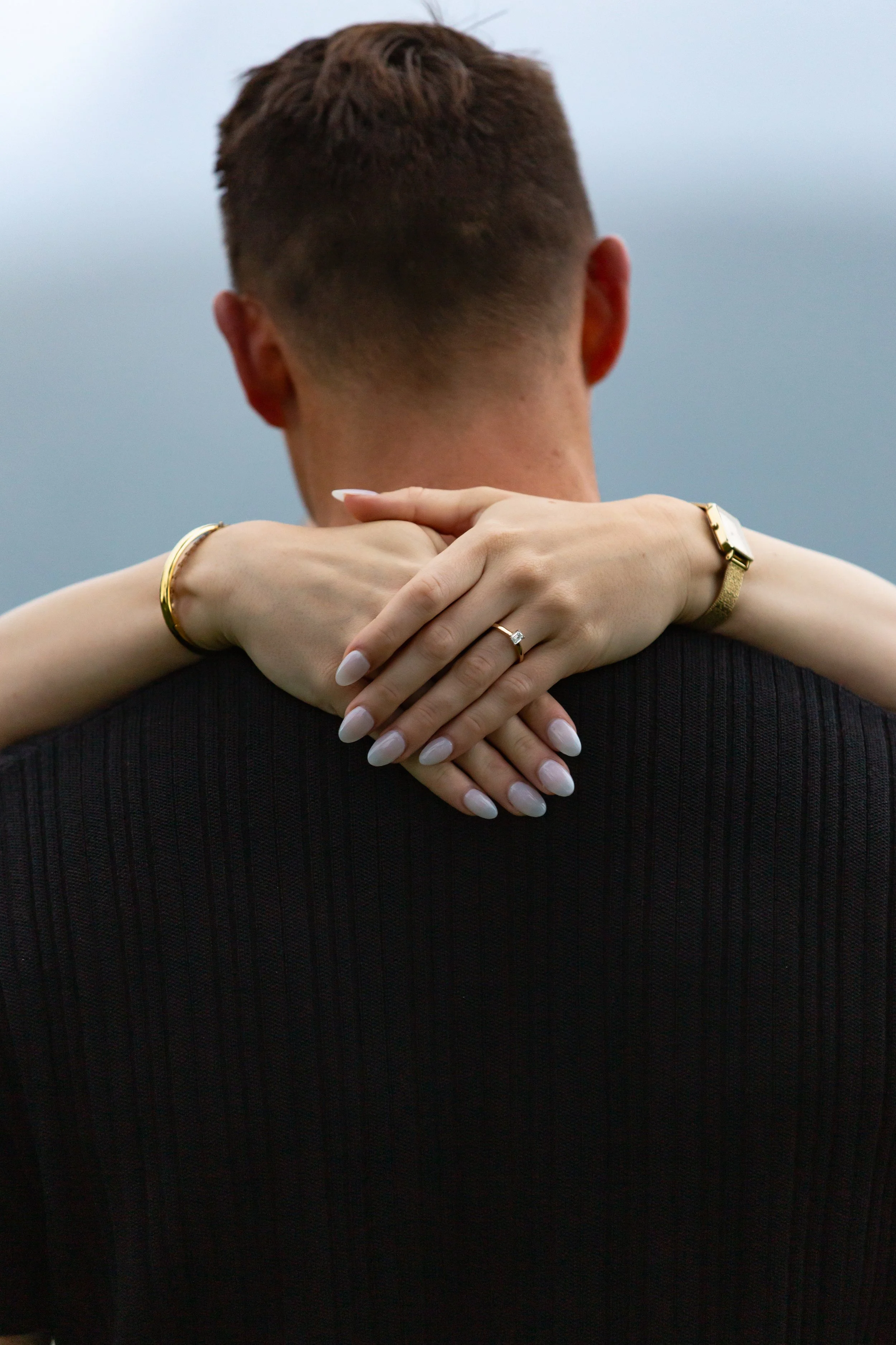 Couple embrace a hug with showing off the new engagement ring in a close up detail.