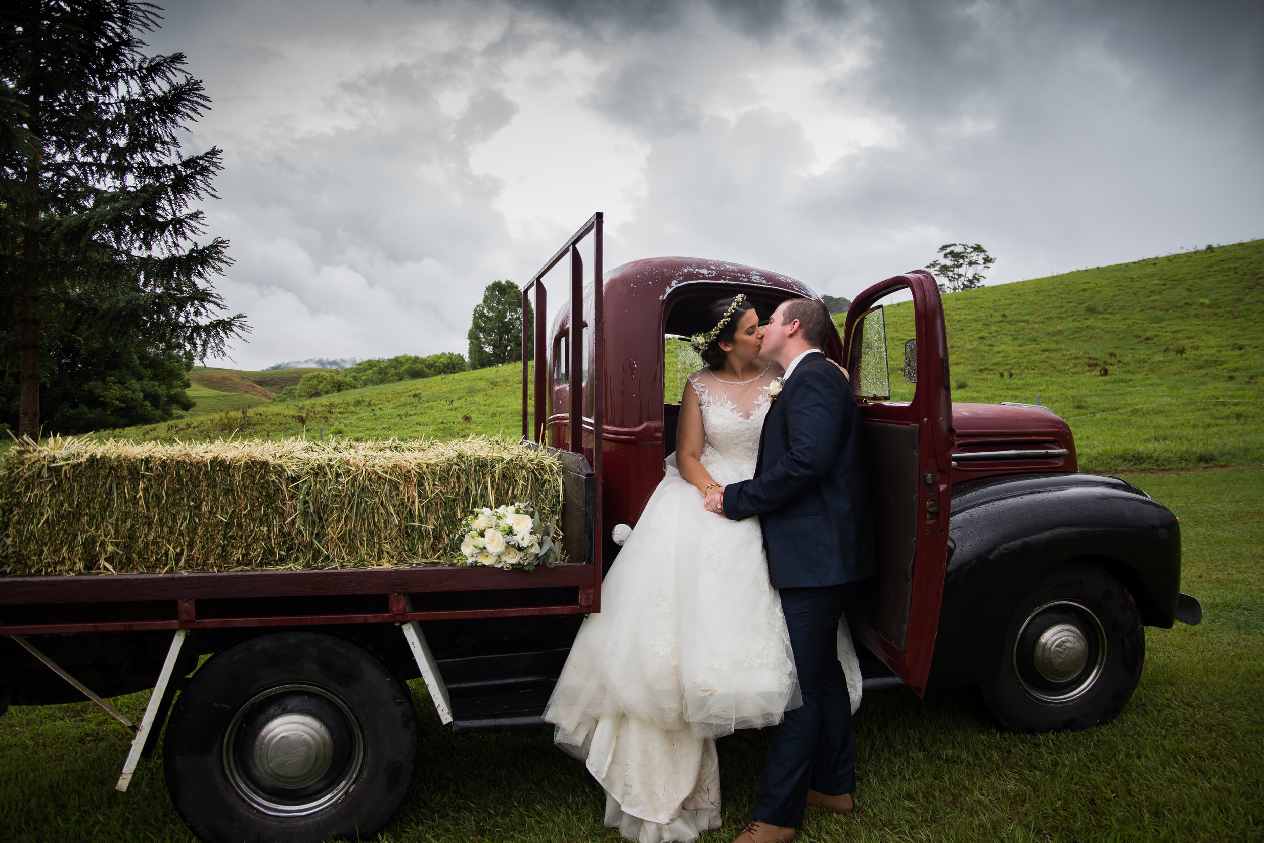 Couple share a kiss in a country farm truck with green hills in the background.