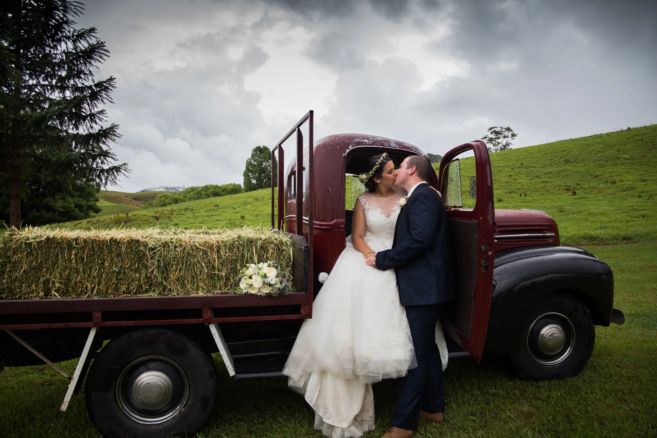 Couple share a kiss in country farm truck with green hills in the background.  