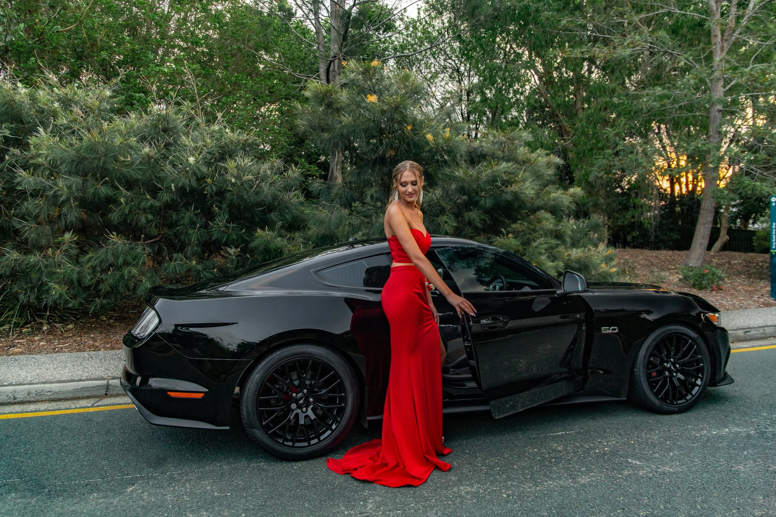 Student going to school formal stands in long red dress beside a Ford Mustang at Benowa Botanical Gardens.