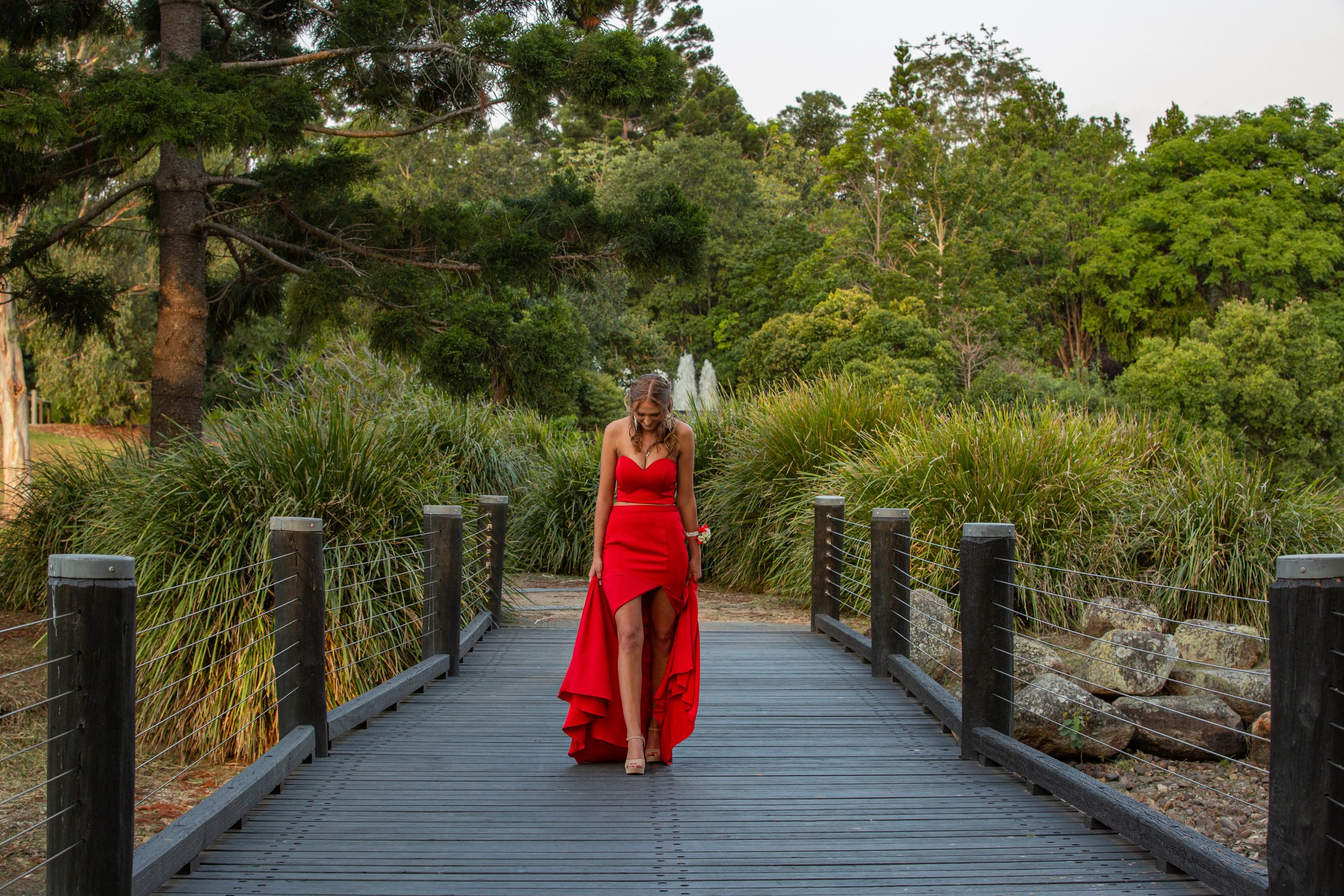Student going to school formal walks in long red dress across bridge during golden sunset at Benowa Botanical Gardens.