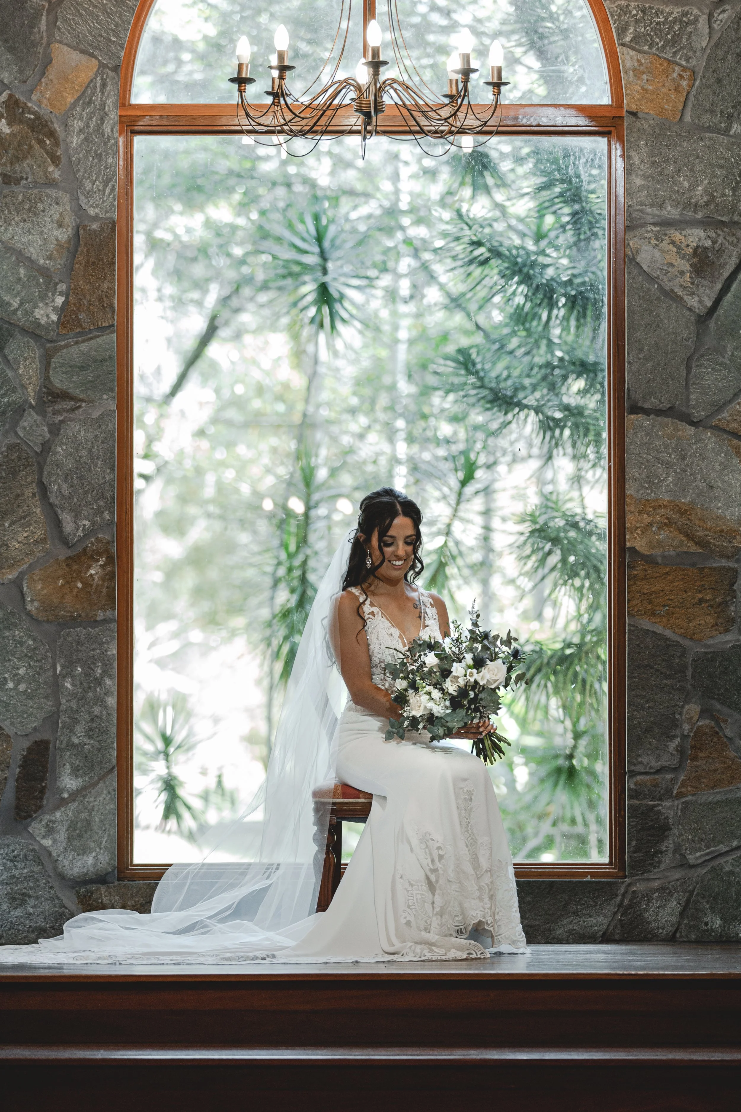 Bride in wedding gown sitting by big arch window holding flowers at Coolabah Downs Private Estate. 
