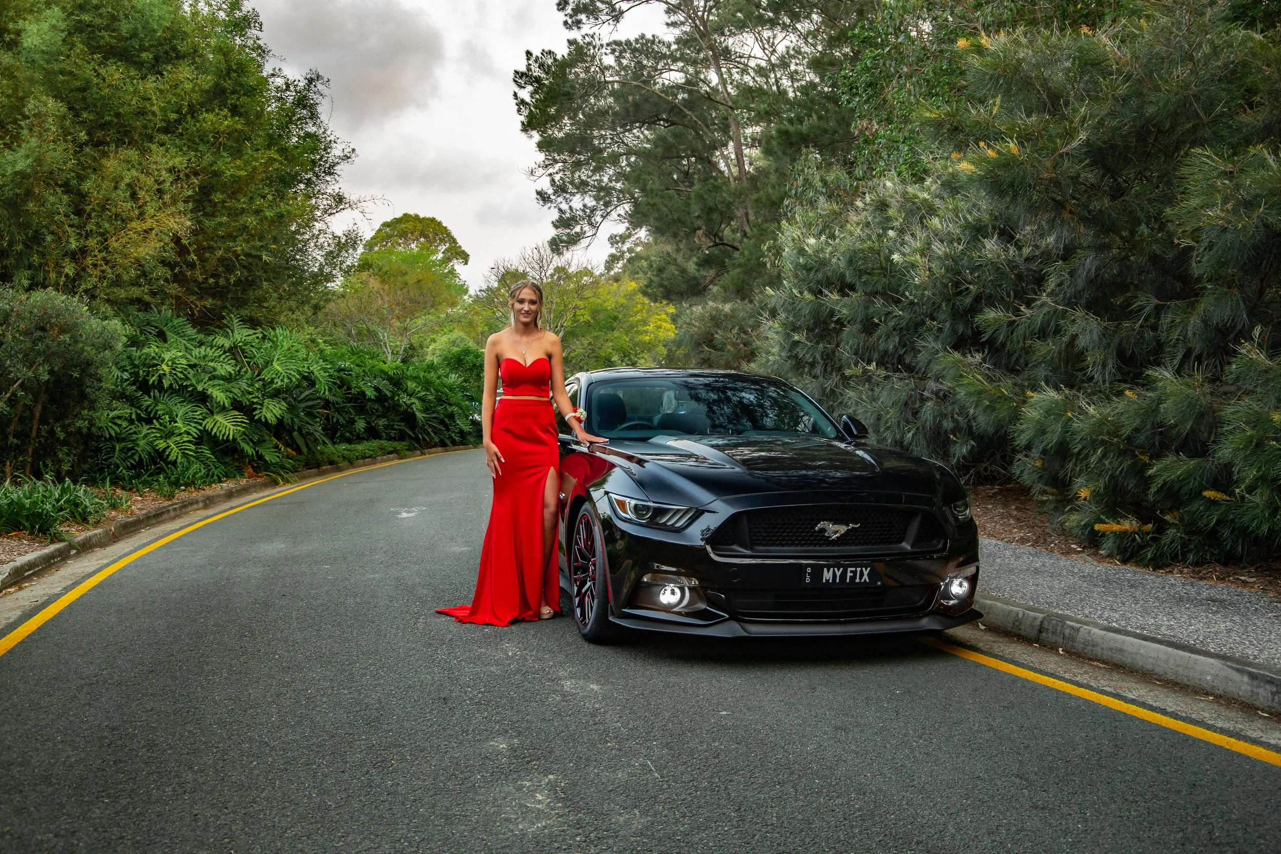Student going to school formal stands in long red dress beside a Ford Mustang at Benowa Botanical Gardens.