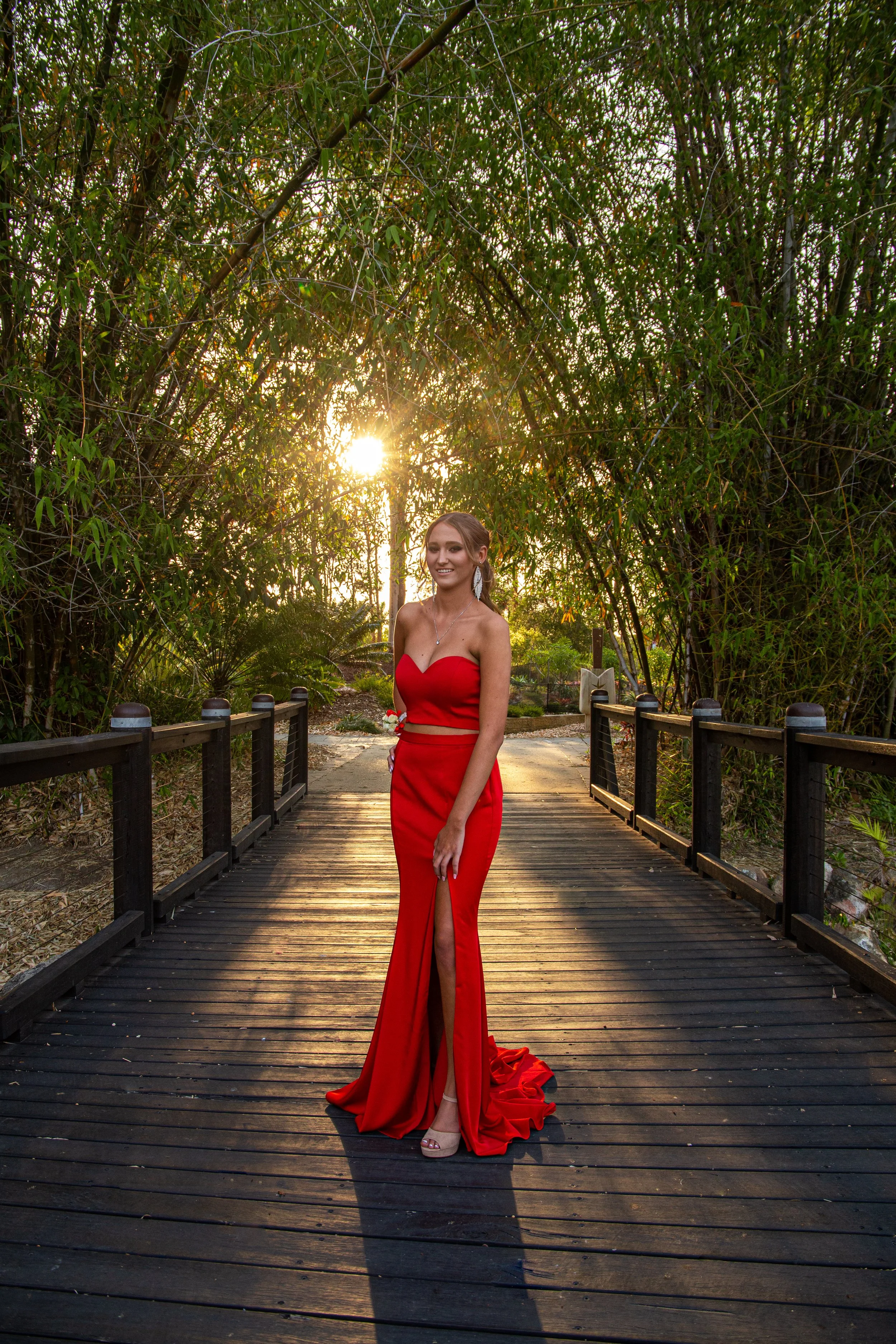 Student going to school formal stands in long red dress on bridge during golden sunset at Benowa Botanical Gardens.