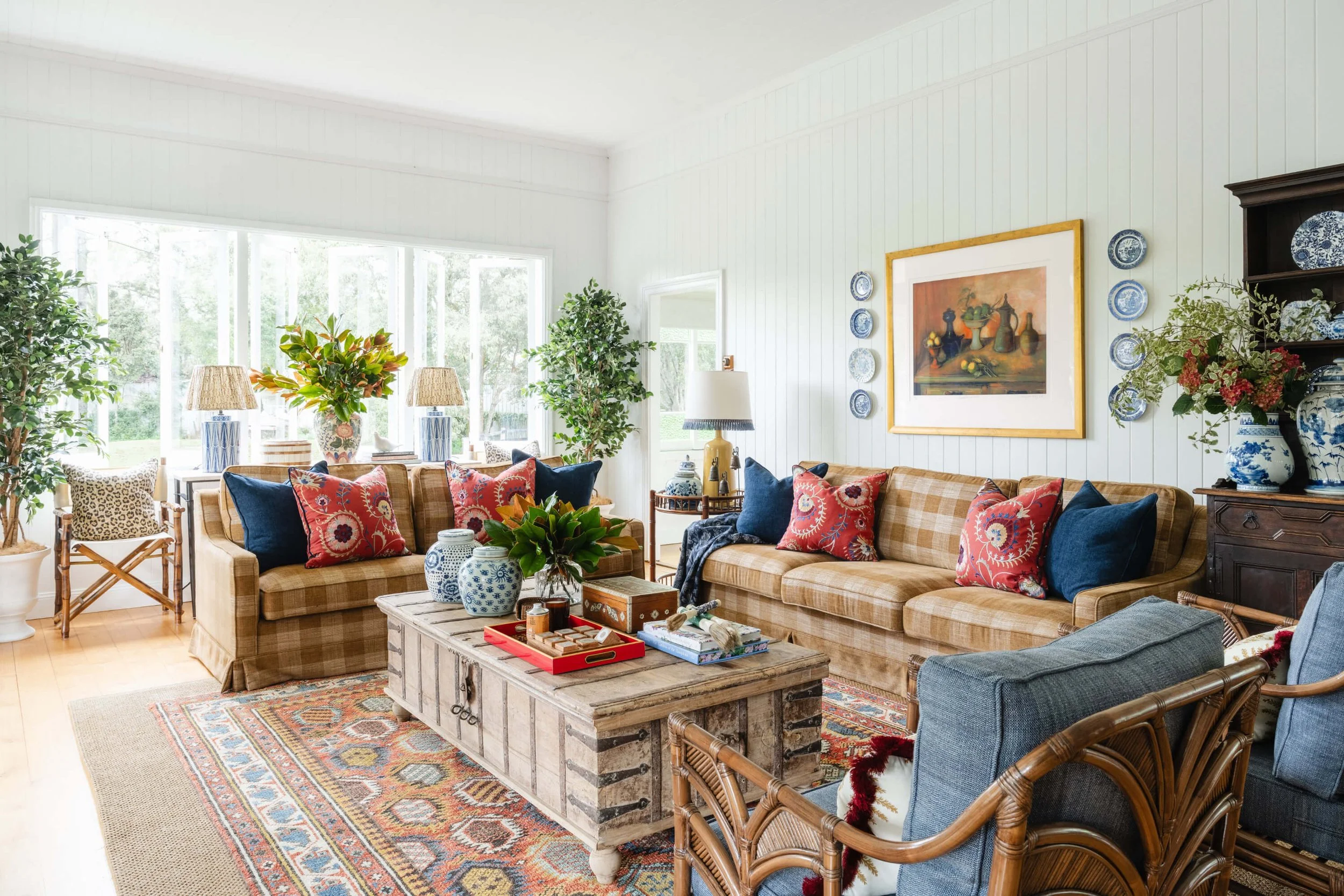 A bright living room with large windows, beige plaid couches with red and blue pillows, a wooden coffee table, potted plants, and a traditional patterned rug. Ascot Residential Interior Design by Biju Interiors.