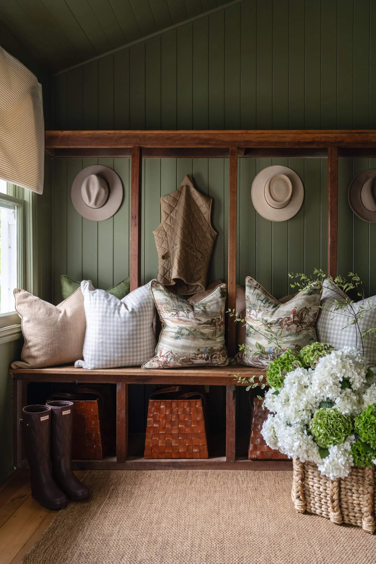 A cozy corner with green wooden wall paneling. Decor includes hats hung on hooks, a quilted vest, and a satchel on wall hooks. Shelves hold decorative items like boots, woven baskets. On the right, a large basket with hydrangeas and green foliage. The bottom shelves contain baskets, and the floor is covered with a textured beige rug.