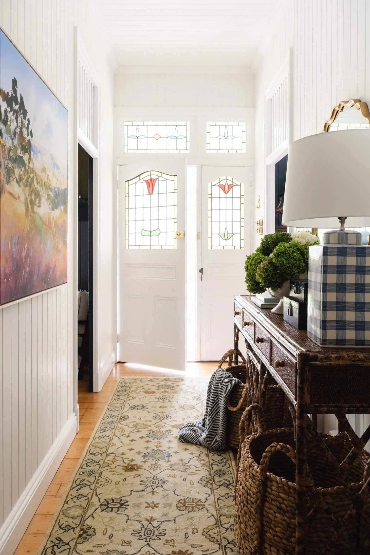 Rosewood Farm entryway or foyer area with a front door featuring decorative stained glass panels and upper windows, a console table with a lamp and decorative greenery, woven baskets, and a light-colored patterned runner rug on a wooden floor.