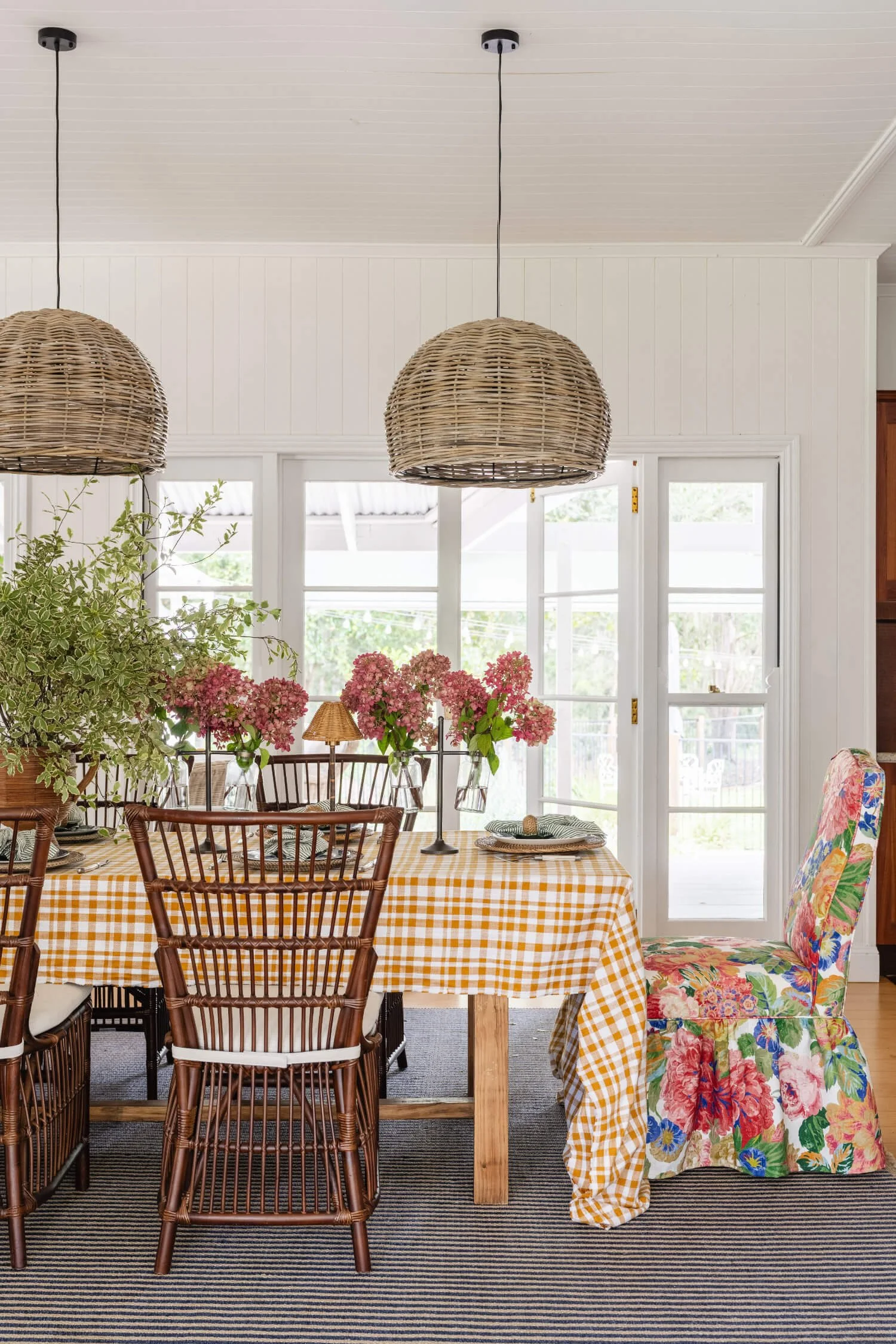 Rosewood Farm dining area with a table covered with a yellow and white checkered tablecloth, surrounded by wooden chairs, with a large floral-patterned armchair on the right. By Biju Interiors.