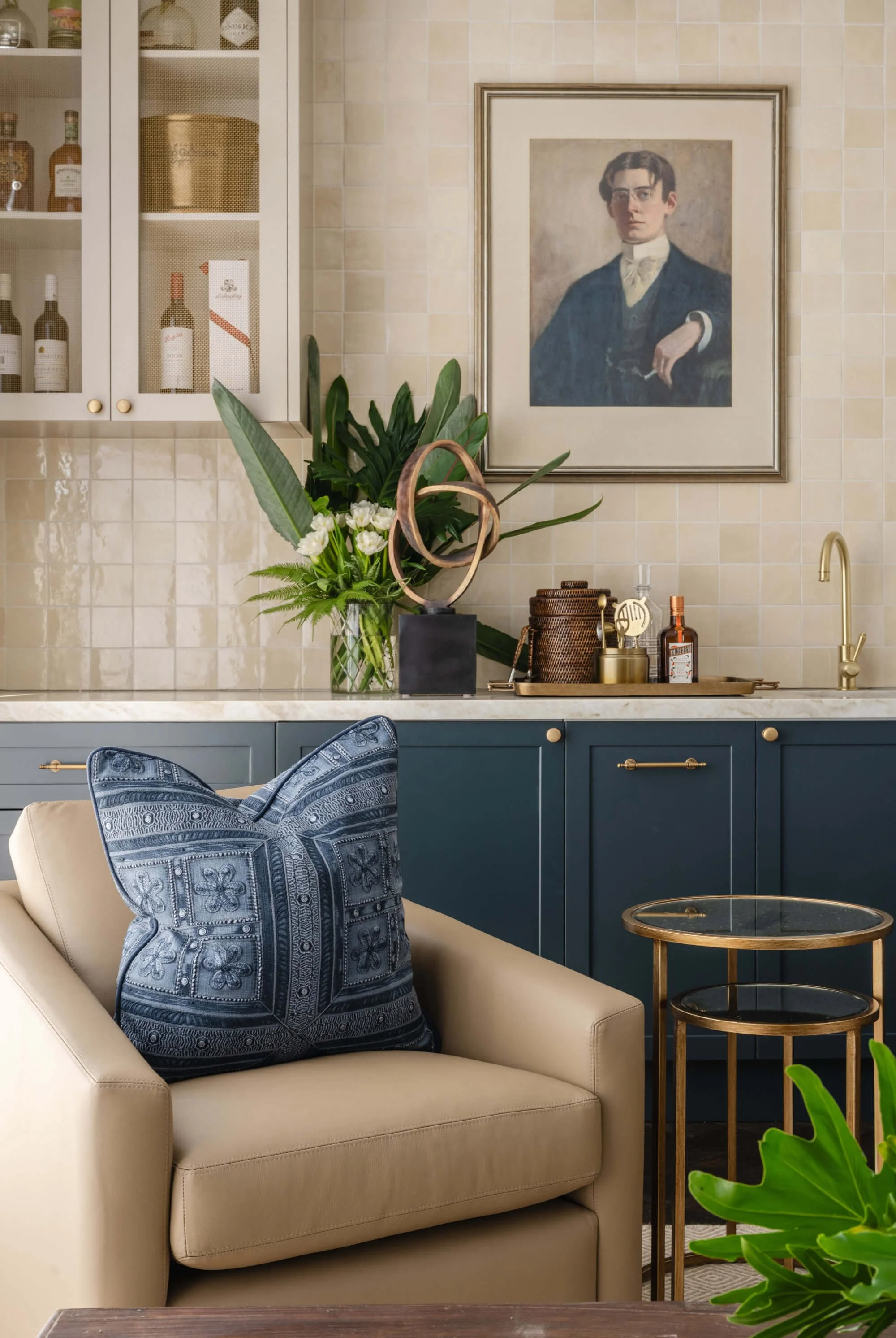 Living room with beige sofa and blue patterned pillow, beige and navy blue cabinetry, a large framed portrait on a beige tiled wall, flowers and greenery on the counter, and gold accents. Ascot Residential Interior Design by Biju Interiors.