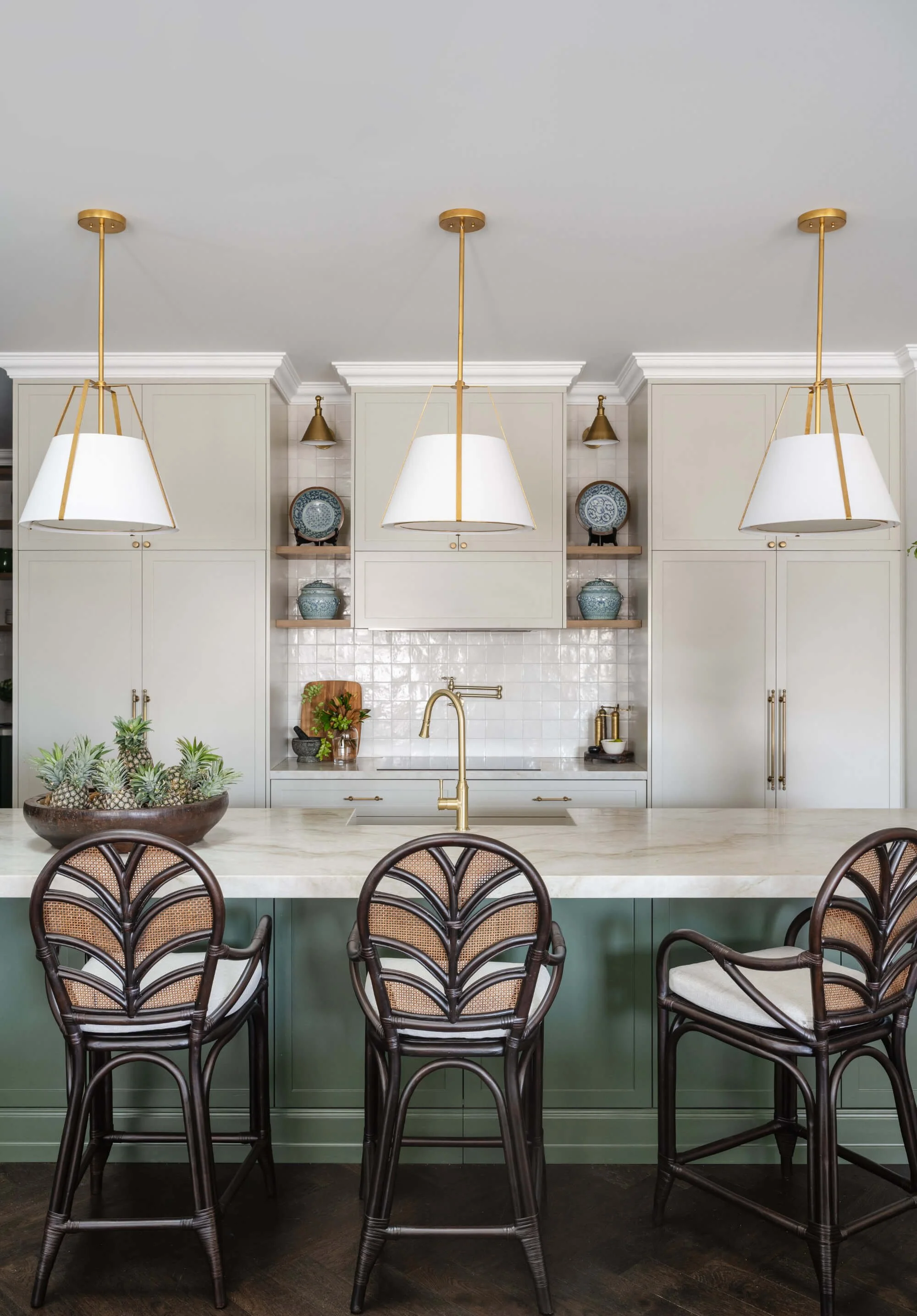 A kitchen with three pendant lights hanging above a marble kitchen island, with dark brown rattan chairs and a black bowl with green succulents on it. Ascot Residential Interior Design by Biju Interiors.