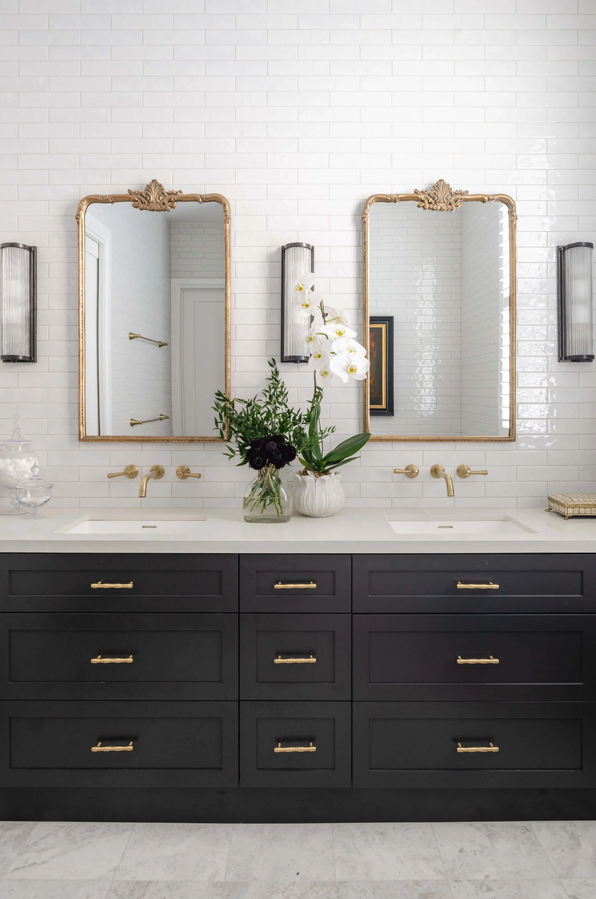 Bathroom vanity with two gold-framed mirrors, black cabinets, gold handles, white countertop, gold fixtures, and a floral arrangement with white orchids and green leaves. Ascot Residential Interior Design by Biju Interiors.