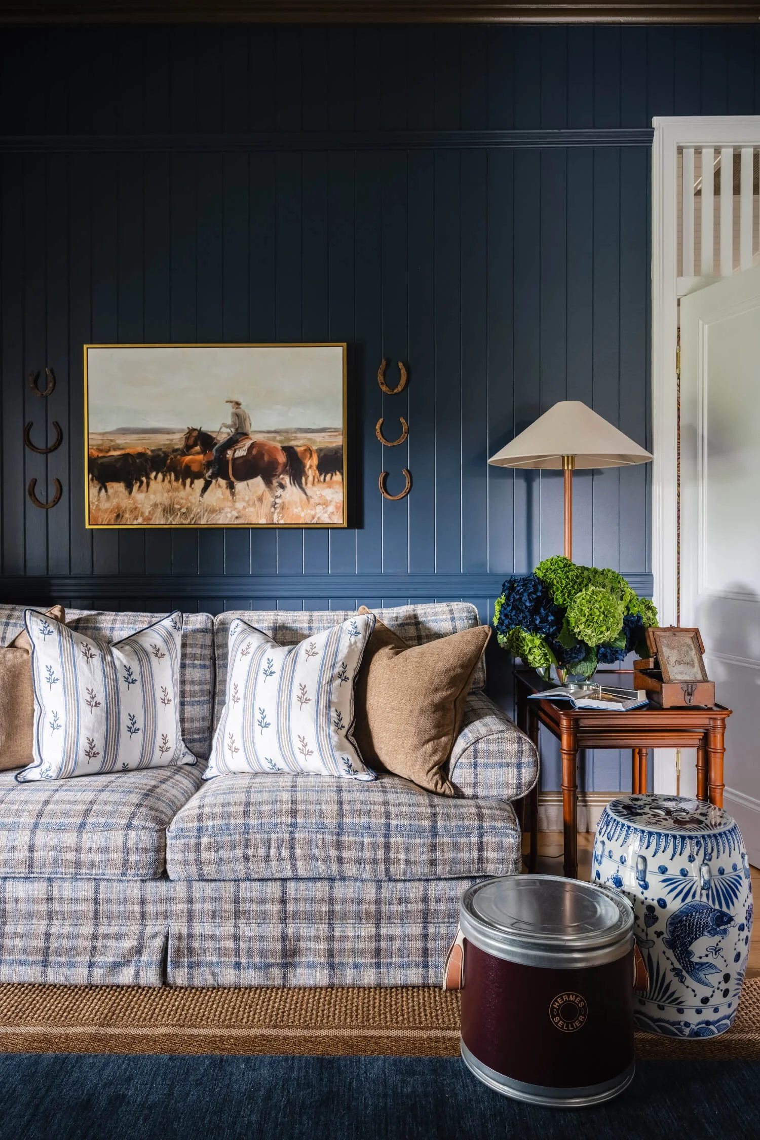 Rosewood Farm living room with a plaid sofa adorned with striped and plain pillows. Behind the sofa, there's a dark blue paneled wall with a landscape painting of a cowboy riding a horse among cattle and horses. By Biju Interiors